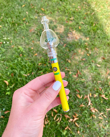 A young Caucasian woman with white fingernails holding a Slim Twist yellow Ooze vape pen with a Cloud Glass Ball attached. She is out on the lawn.