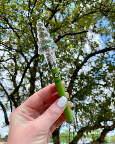 A young Caucasian woman holding a green Ooze Slim Twist vape pen with a Festive Glass Globe in it. She is under the tree and holding a pen against the background of branches and leaves.