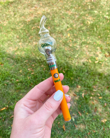 A young woman's hand with white fingernails holds an orange Ooze Slim Twist vape pen with a Swoop Glass Ball attached. She is standing on the grass.