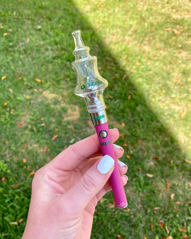 A young woman's hand with white fingernails holds a pink Ooze Slim Twist vape pen with a Genie Glass Ball attached. She is standing on the grass.