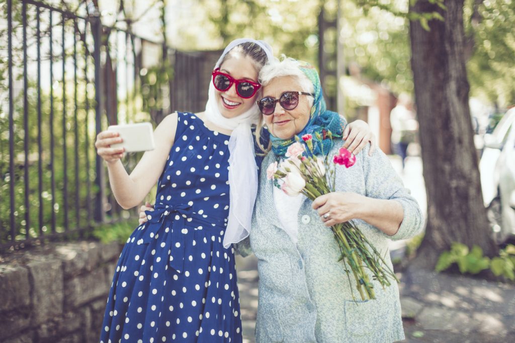 Smiling young girl is taking a selfie of herself and her grandmother while the woman holds a gift and a bouquet of flowers.