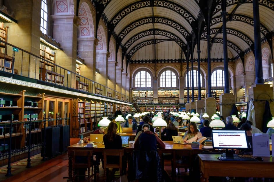 View of the bibliothèque Sainte-Geneviève in Paris. This library, mainly used by university students, is famous for its domes decorated with lace-like metalwork. Below are arched windows and two bookshelves leaning against the wall. In the center of the photo as well as the library are many desks with old-fashioned blue cap lamps beside each seat. Students are sitting working on their computers.
