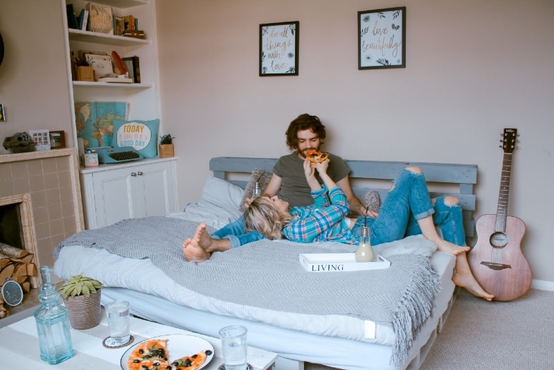 woman feeding man with pizza while lying on bed