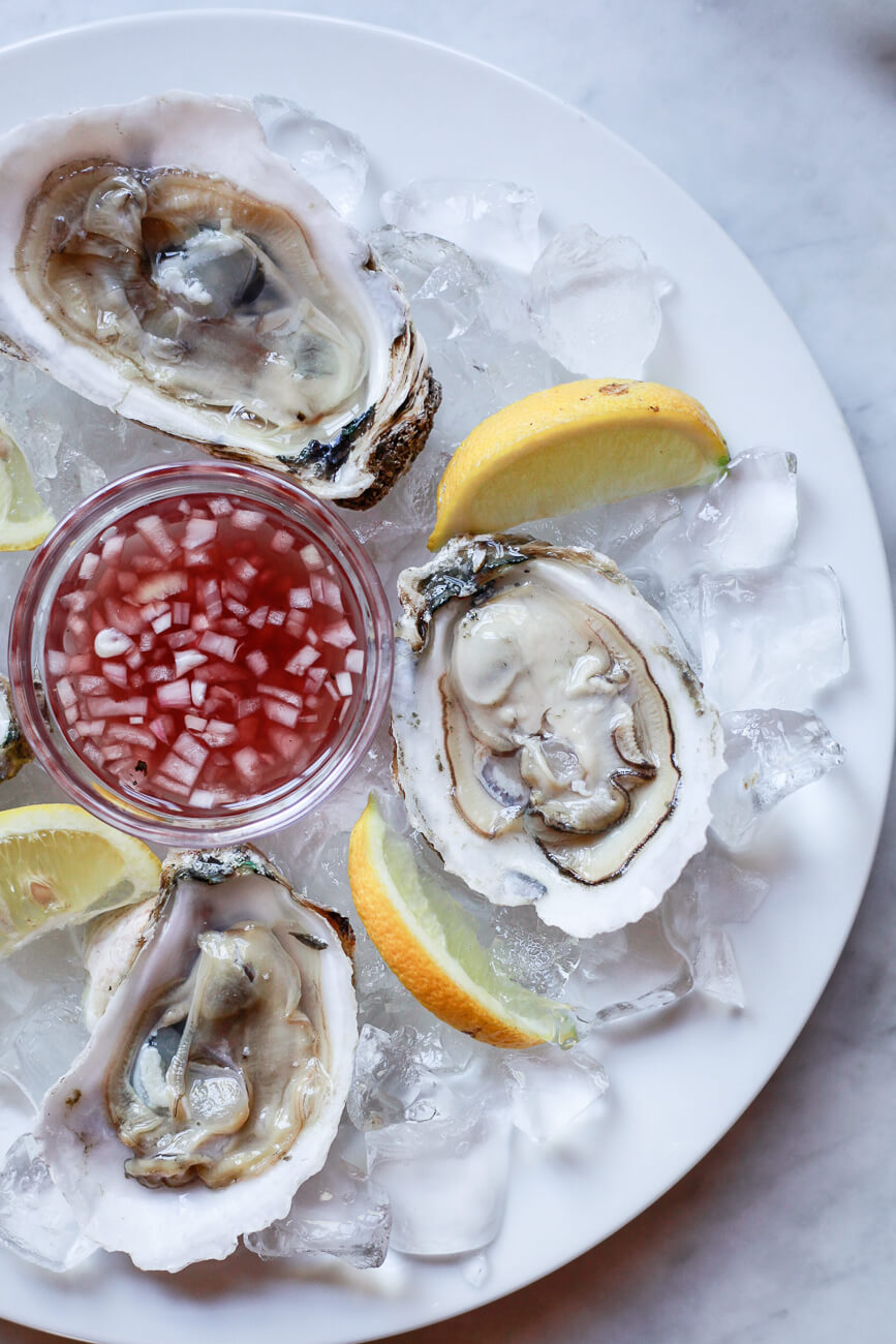 Three raw oysters on the half shell on a plate of ice with a small bowl of mignonette sauce and lemon wedges.