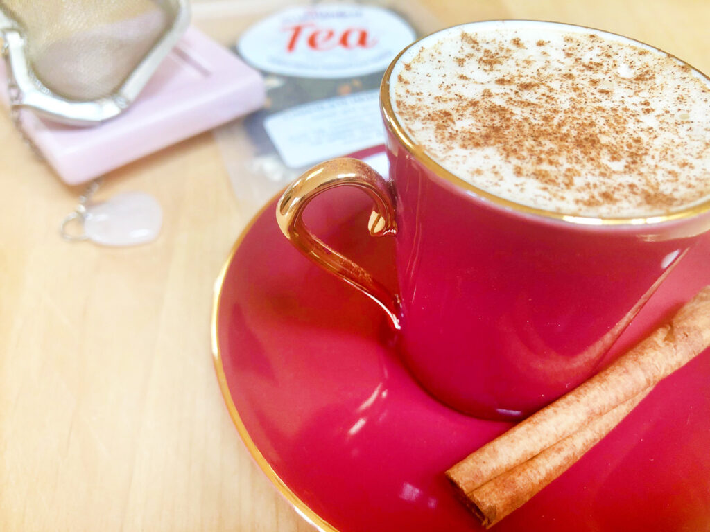 A cup of red tea filled with milk foam and sprinkled with cinnamon rests on the wooden table in front of the heart-shaped mesh tea maker and a loose leaf tea packet. A cinnamon stick placed on a plate.
