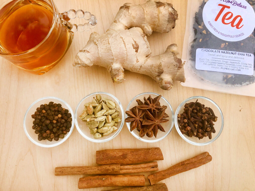 A row of four small bowls filled with pepper, cardamom pods, anise and cloves lay in a row on the wooden table. Beneath them are some cinnamon sticks; Above them was a glass cup full of bottles, a large fresh ginger root, and a packet of loose leaf tea.