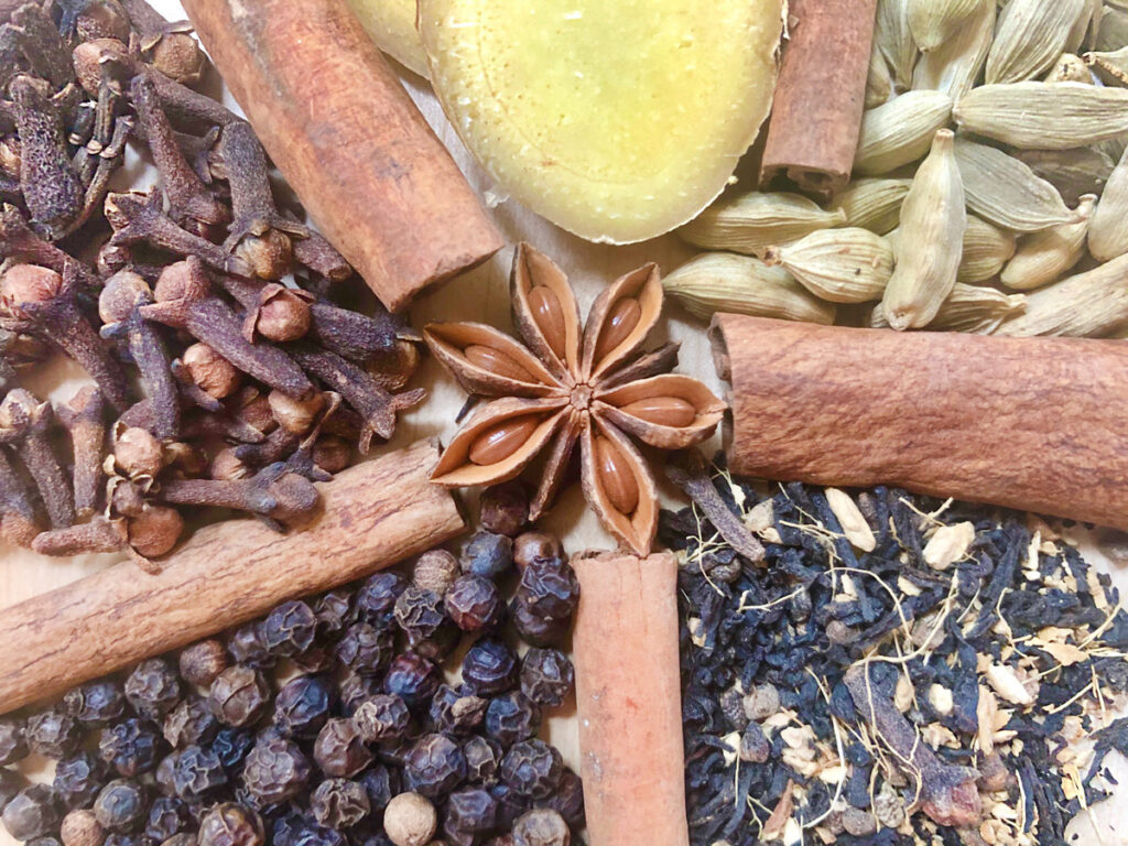 Anise is surrounded by cinnamon sticks, ginger, cardamom pods, peppercorns, cloves and loose leaf chai tea.