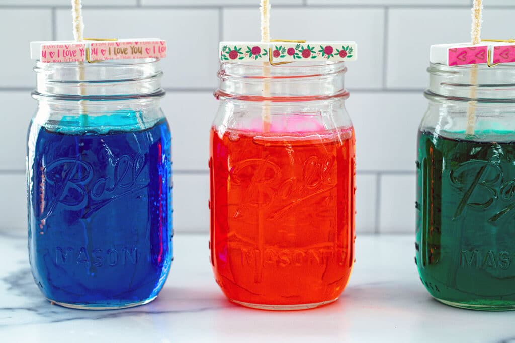 A blue, red, and green jar of sugar solution with sticks hanging out of them for forming rock candy