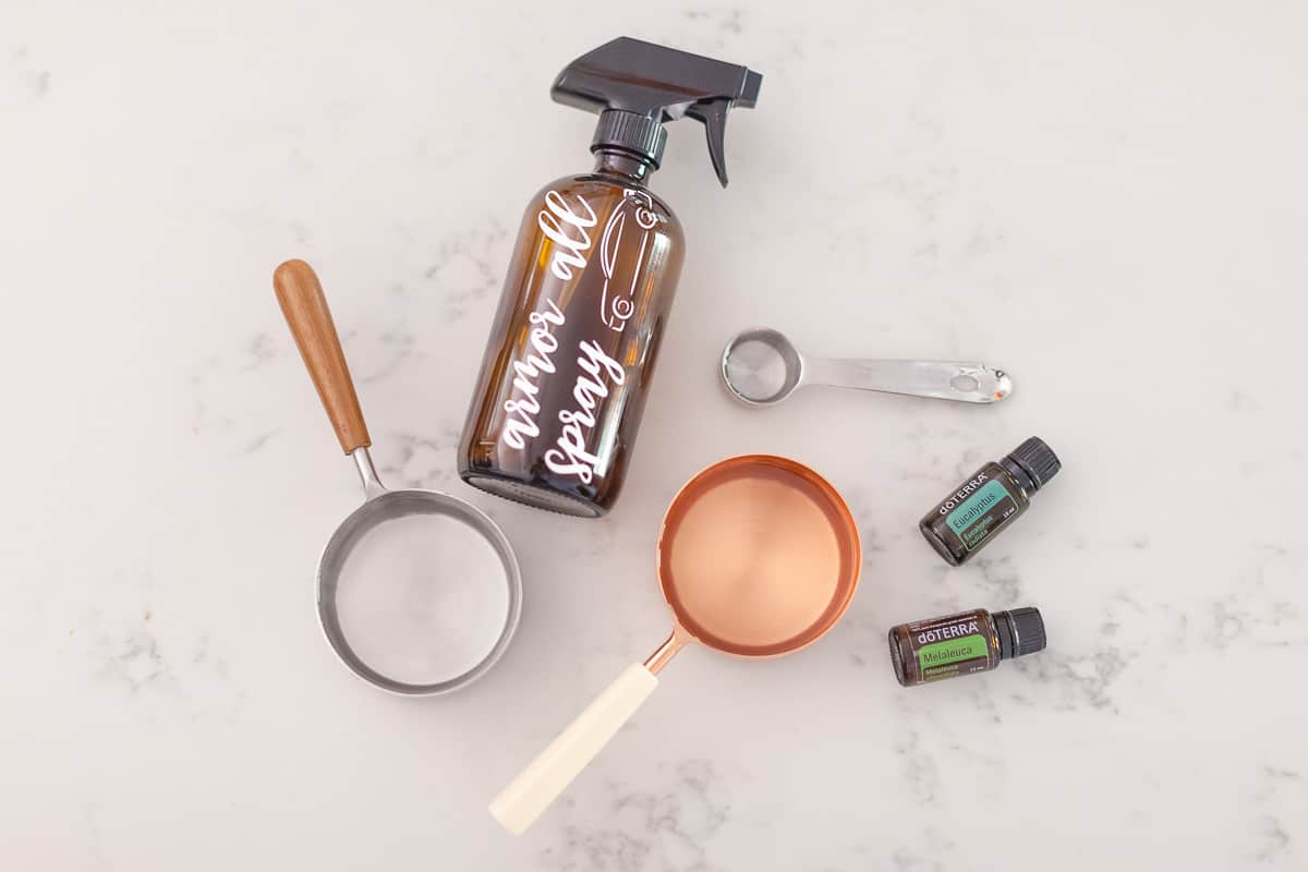There is a silver measuring cup with wooden handle that pours clear liquid into a white funnel, a silver measuring spoon, a copper measuring cup with a white handle, a bottle of eucalyptus essential oil, a bottle of doterra cajeput essential oil and a spray bottle head.