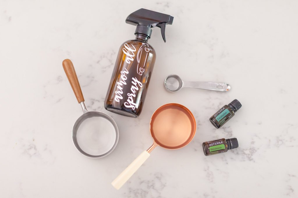 Features a brass measuring cup with white handle for pouring clear liquid into a white funnel, a silver measuring spoon, a bottle of Eucalyptus oil, a bottle of doterra cajeput oil and a spray bottle cap.