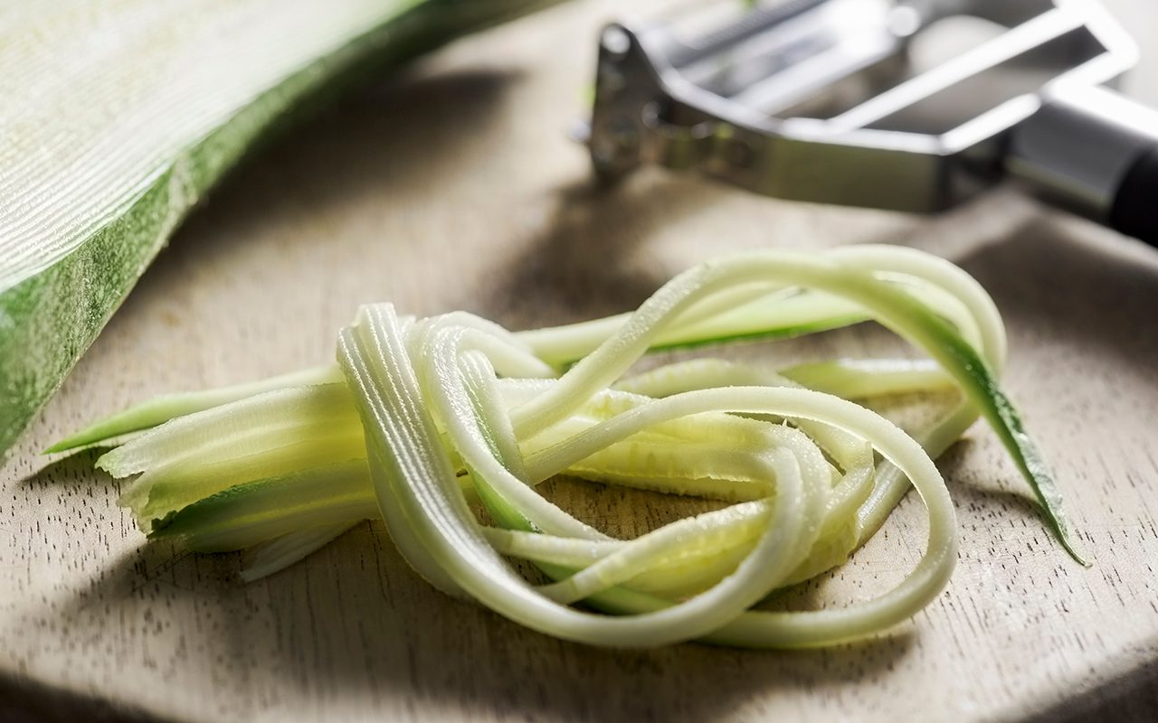 how to grate zucchini The fibers of a zucchini are lying in front of a wooden cutting board. Julienne's belongings and the rest of the household utensils are behind it. The peeler is made from stainless steel and has a black handle. The focus is selectively on the graceful object in front.