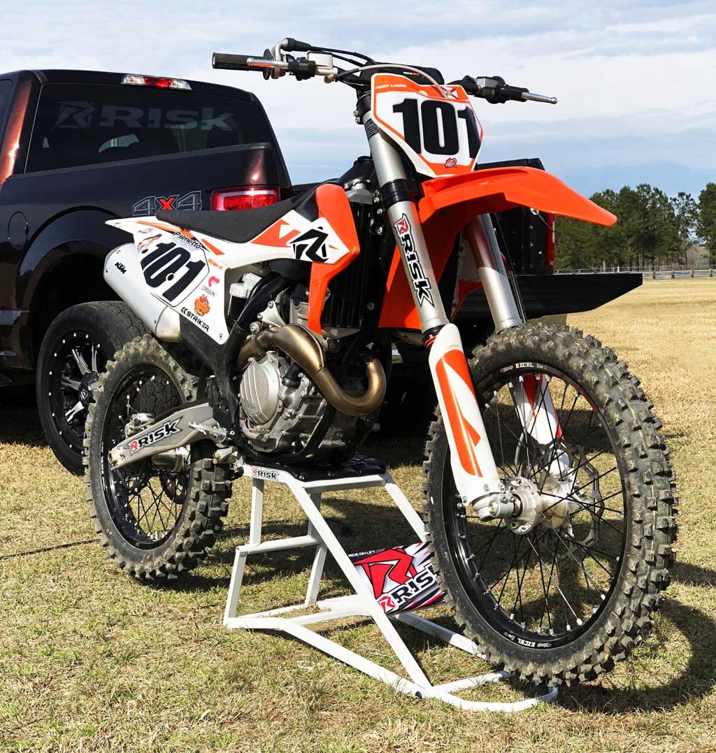 101 motocross dirt bike sitting on a rr1 lift stand by Risk Racing in front of big black truck with Risk Racing window sticker