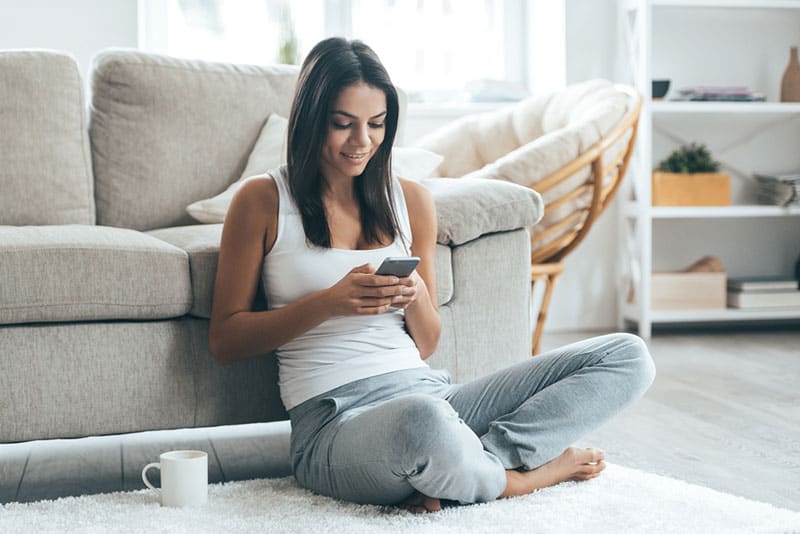 Lovely message from him. Attractive young woman looking at her smart phone and smiling while sitting on the carpet at home