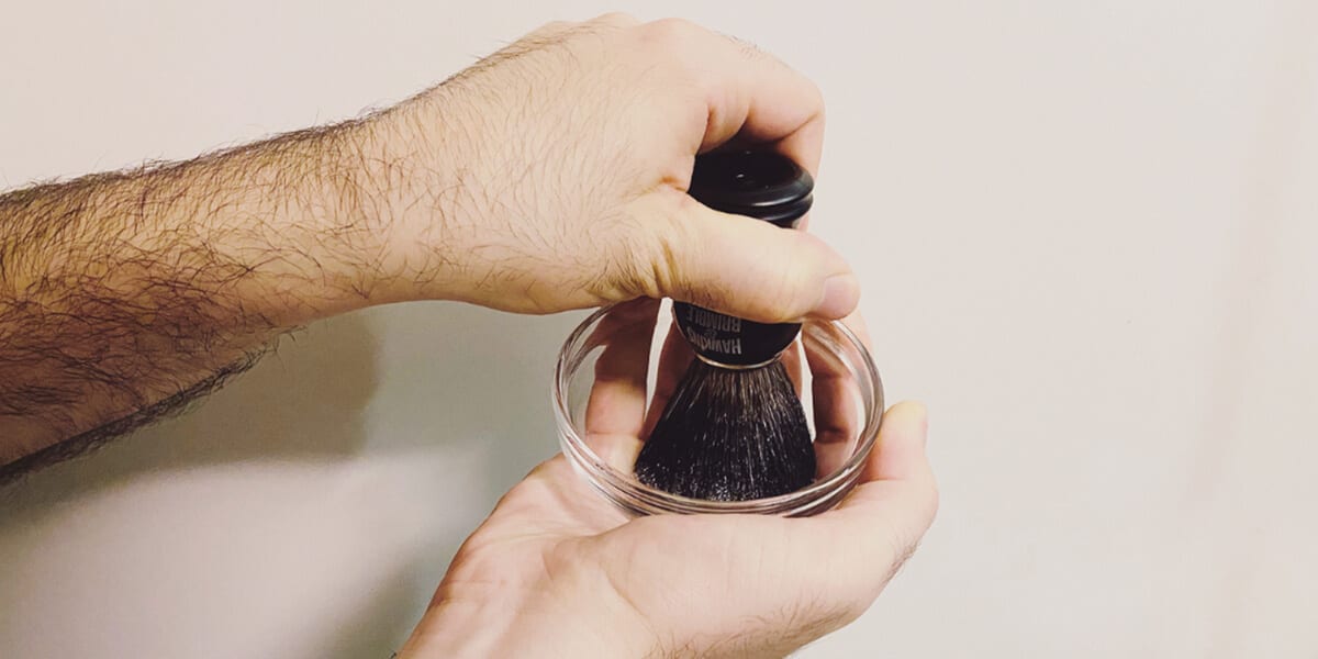 a hand holding beard brush and lathering bristles in bowl of shaving cream